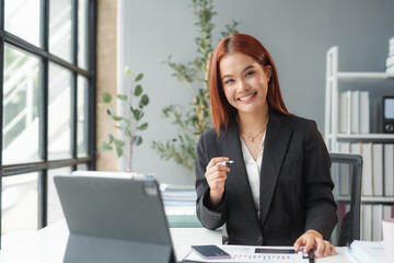 Wall Mural - Young asian businesswoman smiling while sitting at her desk in a modern office, working on a laptop computer with paperwork and financial charts