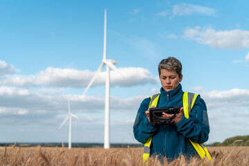 Wind energy technician operates drone in wheat field under bright blue sky with wind turbines spinning in background