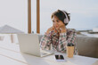© ViDi Studio - Young IT woman wear shirt casual clothes sits alone at street cafe table in coffee shop working or study on laptop pc computer listen music in headphones rest in restaurant during free time outdoor