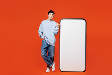 Wall Mural - Full body young man wears blue sweatshirt casual clothes point finger on big huge blank screen mobile cell phone smartphone with area isolated on plain red orange color background. Lifestyle concept.