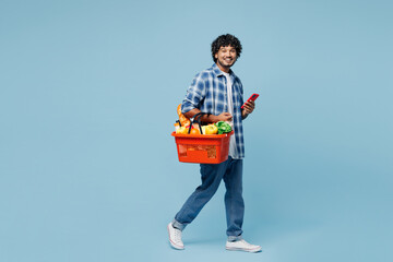 Sticker - Full body sideways young Indian man wear shirt hold red basket with food products vegetables use mobile cell phone isolated on plain pastel blue background. Delivery service from shop or restaurant.