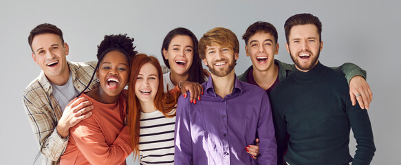 Happy diverse friends having fun together. Banner with group photo of seven cheerful multiracial people in casual clothes standing by grey studio background, hugging, looking at camera and smiling