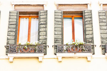 facade of a Venetian building with two windows decorated with peeling green shutters and elegant metal balconies with flower pots. The wooden framed windows, filled with soft daylight, highlight the c