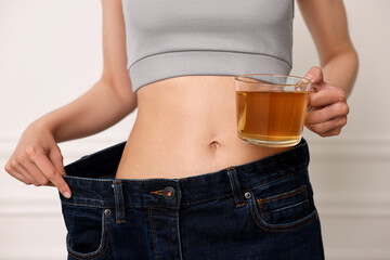 Wall Mural - Weight loss concept. Woman with cup of diet tea and big pants near white wall, closeup