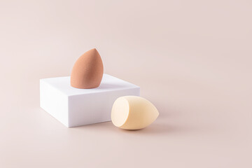 Two cosmetic egg-shaped blender sponges on a white square catwalk. Beige background. Front view. Care and beauty concept. Makeup tool.