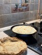 © Anastasia - A cozy kitchen photo with a focus on making crepes. A stovetop with a black frying pan containing a cooking crepe, positioned on a lit gas burner. In the foreground, there is a stack of cooked crepes.