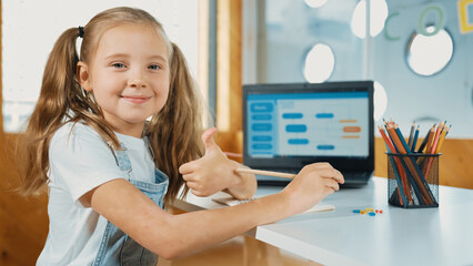 Little pretty caucasian girl smiling to camera while showing a thumb up gesture. Young child wearing headphone and casual dress while laptop display coding system or programing prompt. Erudition.