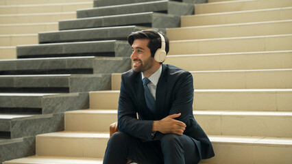 Wall Mural - Project manager listening and enjoy music from headphone while sitting at stairs. Professional businessman wearing suit outfit while dancing and moving to music. Happy man listen funny song. Exultant.