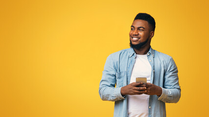 Wall Mural - Handsome black man holding smartphone and looking aside at copy space on yellow studio background