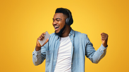 Poster - Joyful african american man in wireless headphones singing into smartphone like microphone on yellow background, panorama