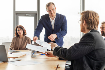 Aufkleber - A businessman stands at the head of a conference table, distributing papers to a colleague seated at the table. Other colleagues sit at the table, listening to the businessman.