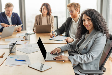 Aufkleber - A woman with curly hair sits at a conference table and types on her laptop while other business professionals work around her. A large window with a city view is visible in the background.