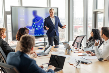 Poster - A businessman stands in front of a large screen displaying a financial chart, pointing to the data and presenting it to a group of colleagues in a modern office.