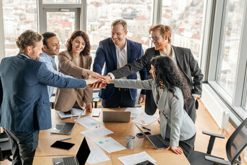 Aufkleber - A diverse team of business professionals stand around a conference table, their hands stacked in the center. They smile at each other, showcasing their positive and collaborative work environment.