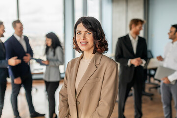 Aufkleber - A businesswoman in a beige blazer stands confidently in the foreground of a modern office setting, smiling at the camera while colleagues work in the background.