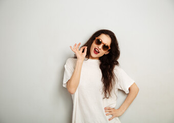 Wall Mural - Fun playful woman posing in fashion sunglasses and white t-shirt and showing okey sign by fingers on white wall background with empty copy space. Happy lifestyle