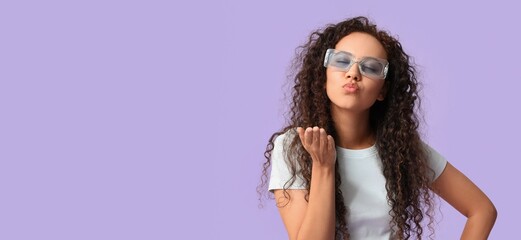 Canvas Print - Beautiful African-American woman with sunglasses blowing kiss on lilac background with space for text