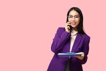 Wall Mural - Portrait of young Asian businesswoman with clipboard talking by phone on pink background