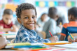 © Marina Varnava - Happy boy writing with a red crayon at a classroom desk surrounded by peers engaged in activities