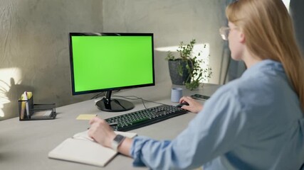 Young Woman Looks at Computer with Green Screen Mock Up Display and Writes Something in Notebook. Girl Working In Office On Computer With Green Screen