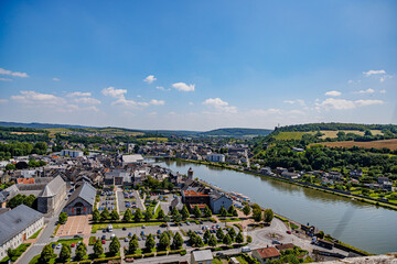 Aerial view of Meuse river and Givet city in panoramic view, buildings, parking lot, streets, houses, trees on hill and skyline in background, sunny summer day in Ardennes department, France