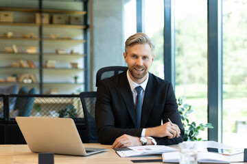 Portrait of happy businessman sitting at office desk, looking at camera, smiling.