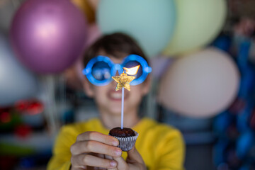 Cute little boy in a yellow T-shirt and blue party glasses holding birthday cupcake with candle 