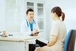 © Phushutter - A male Asian pediatrician in a white lab coat sits at his desk, warmly greeting a middle-aged pregnant woman for a prenatal consultation, offering care and support in a clinical setting.
