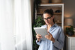 © Srdjan - Real estate agent with tablet pc looking through window. Young business woman working at home, using device gadget. Successful professional occupation graphic designer female taking a break from work.