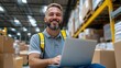 © Pinklife - A cheerful warehouse worker with a bearded smile wears a reflective yellow vest and sits with a laptop amidst packages. Stacks of boxes form the warehouse backdrop.