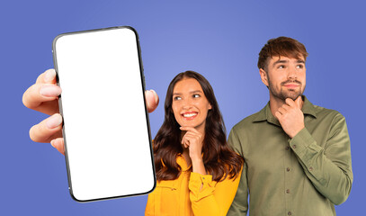 Fototapete - A woman holds a smartphone with a blank screen in front of a blue backdrop. She is smiling and looking at the screen. The man beside her is looking upwards while holding his chin