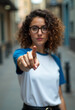 © Limanou Mikael - A young woman with curly hair, wearing glasses, points her finger in front of her on the street.