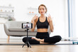 © sofiko14 - Yoga instructor sitting cross-legged on mat recording virtual fitness class using smartphone. Instructor demonstrating poses in modern living room. Concept of online fitness and digital wellness.