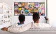 © Prostock-studio - Two men are relaxing on a couch in a living room, watching television. The television is displaying a large, colorful grid of images.