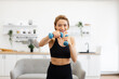 © sofiko14 - Energetic woman in workout attire exercising with dumbbells at home in bright modern living room. Concept of fitness, health, and home workout routines.