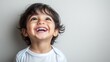 © mashimara - Happy toddler boy with a big smile, showing his teeth, looking upwards with joy, against a light background.