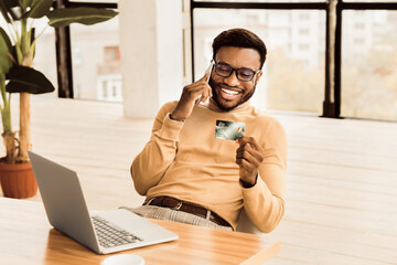 Poster - Easy Payments. Happy african worker holding credit card after online purchase and talking on mobile phone. Copy space