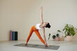 © DragonImages - Woman practicing yoga stretching on yoga mat in living room with plants and exercise equipment. She is in yoga pose looking upwards with arm extended