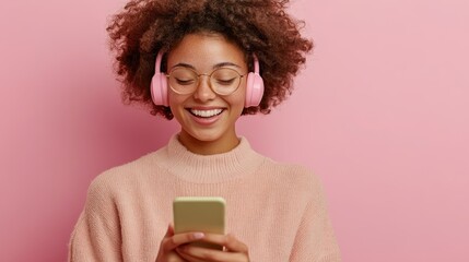 Wall Mural - Young woman with glasses and curly hair, wearing a pale pink sweater and pink headphones, grins at her phone, showcasing a moment of digital delight and relaxation.