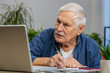 © Andrii Iemelianenko - Senior man accounting payments calculating monthly expenses bank bills analyzing paperwork at home apartment indoors. Elderly mature grandfather freelancer working online remote job on laptop at desk
