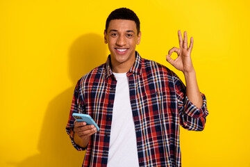 Wall Mural - Portrait of nice young guy hold smart phone show okey symbol wear shirt isolated on yellow color background