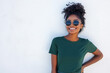 © GustavsMD - Smiling woman in sunglasses and green t-shirt standing against a white wall. Outdoor fashion portrait. Summer style concept