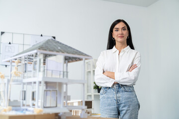 Close up portrait of young beautiful professional architect engineer crosses arms and poses confidently with house model at white modern office with blueprint. Creative living and design. Immaculate.