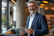 © mankjon - A confident middle-aged man with gray hair and a beard wearing a navy blue suit and glasses, holding a tablet, smiling warmly in a modern office with soft lighting and blurred background