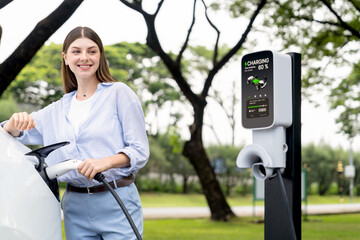 Wall Mural - Young woman recharge EV electric vehicle's battery from EV charging station in outdoor green city park scenic. Eco friendly urban transport and commute with eco friendly EV car travel. Exalt