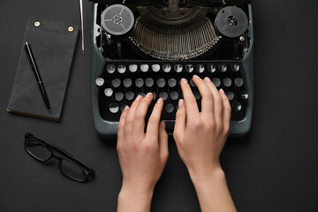 Wall Mural - Woman typing on typewriter with eyeglasses, notebook and pen on black background