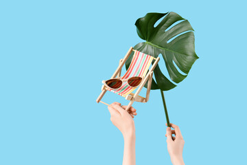 Wall Mural - Female hands with sunglasses, mini deck chair and monstera leaf on blue background. Travel concept