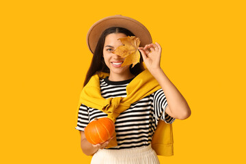 Wall Mural - Stylish young woman with fresh pumpkin and autumn leaf on yellow background