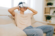 © SHOTPRIME STUDIO - Virtual reality experience, young man enjoying VR headset while sitting on a sofa, indoor setting with natural light and soft colors, relaxed atmosphere