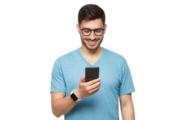 Young handsome man in blue t-shirt holding smartphone and looking at phone screen with smile while standing